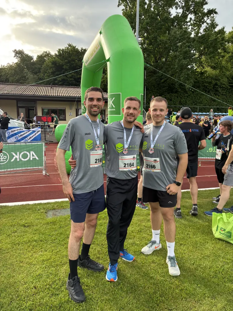 Three runners wearing matching grey ECKERLE E-Motion Technologies shirts, race bibs, and medals pose in front of a green inflatable race arch.