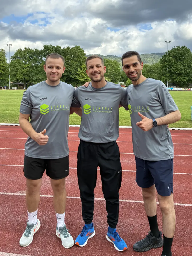 Three runners in matching grey ECKERLE E-Motion Technologies shirts pose together on an outdoor running track.